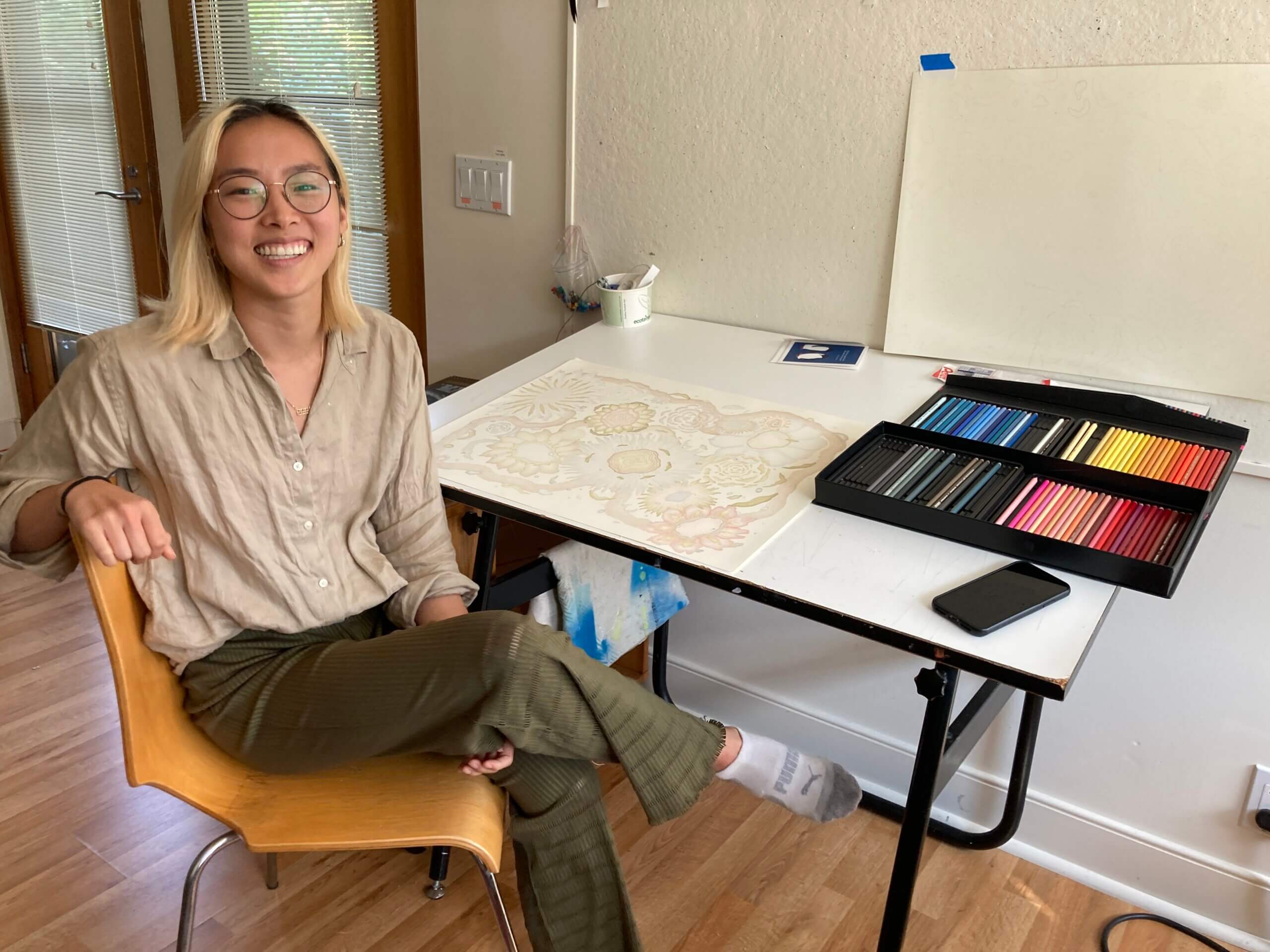 Grace Kim, smiling, sitting at a desk filled with colored pencils and a large work-in-progress