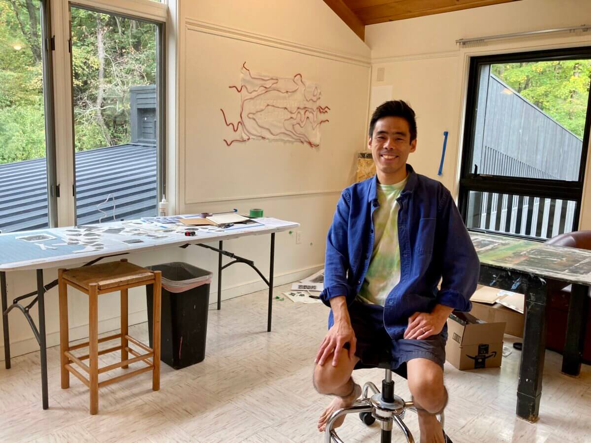 artist Frank Chang sitting in his Saltonstall studio with works-in-progress on tables and walls around him