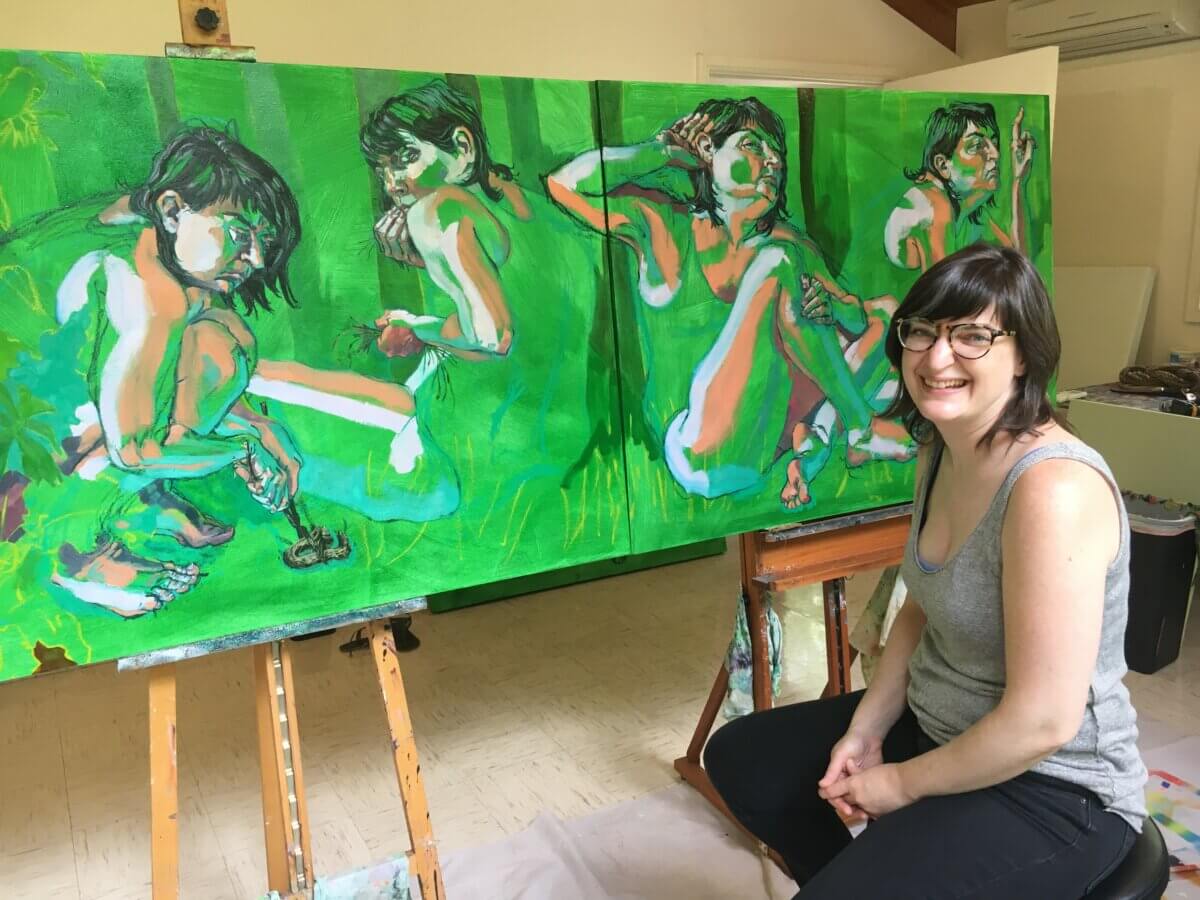 artist Dara Engler sitting in front of a wide multi-paneled painting of a female figure in the woods