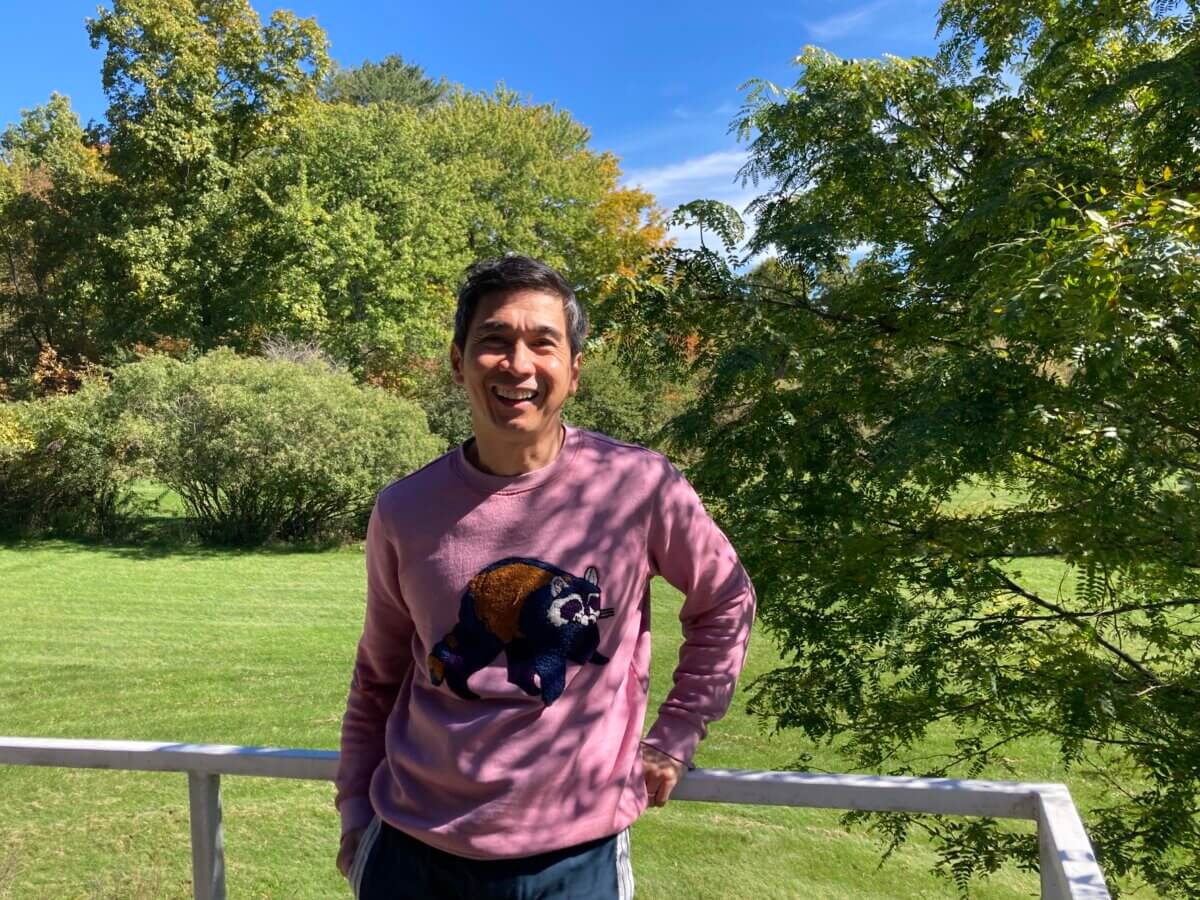 poet Joseph O. Legaspi standing on a balcony in dappled sunlight with green lawn and trees behind him