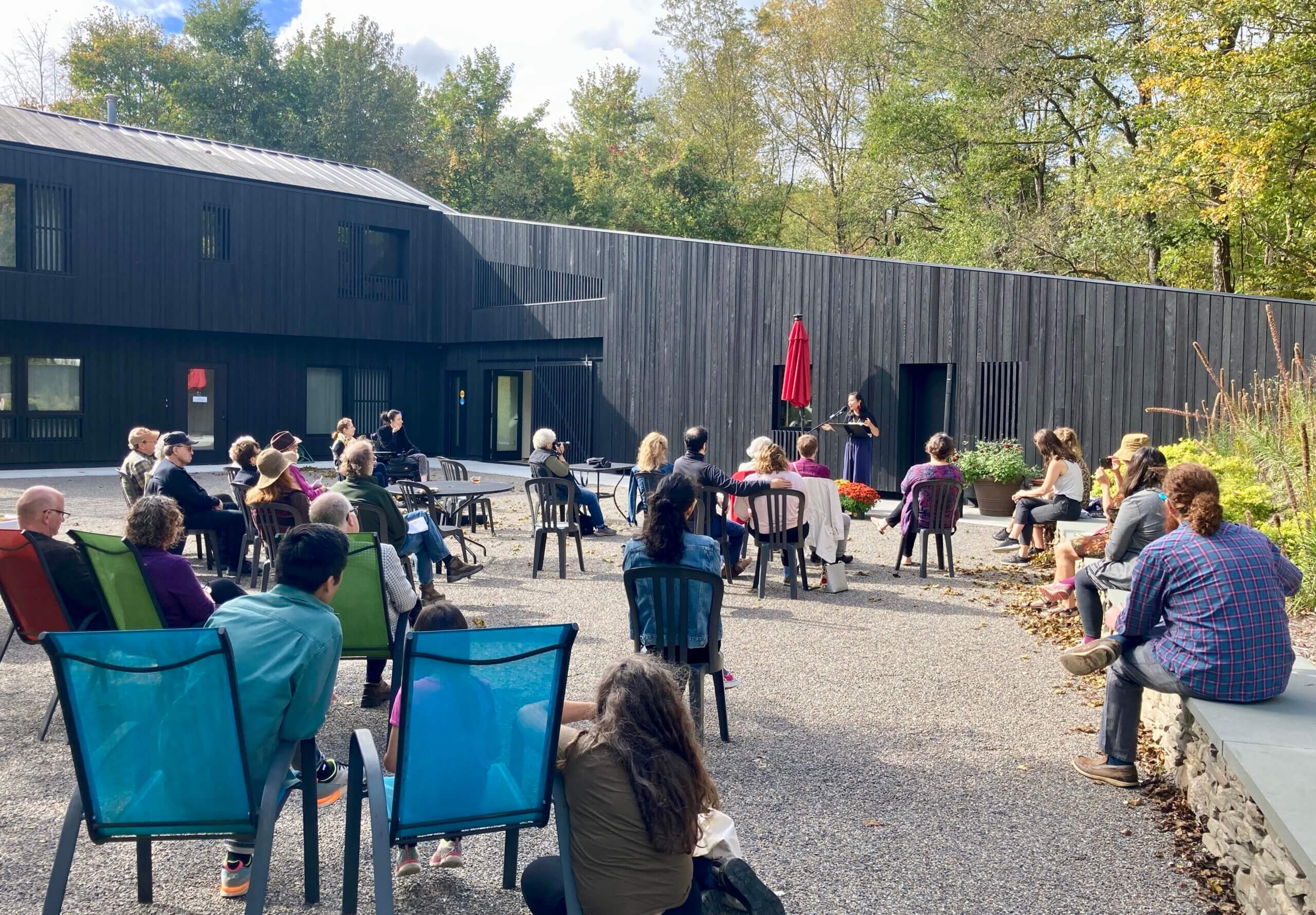 Cathy Linh Che reading at Open House 2021 Asian American poet Cathy Linh Che is standing in front of a mic and music stand, reading to approximately 25 people seated outside on a gravel courtyard. Saltonstall's new black-sided addition is directly behind Cathy. It is early fall, and there is some color in the trees behind the building.